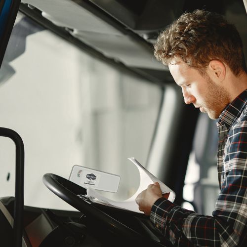 A trucker wearing plaid is sitting in the driver's seat, a clipboard against his steering wheel.