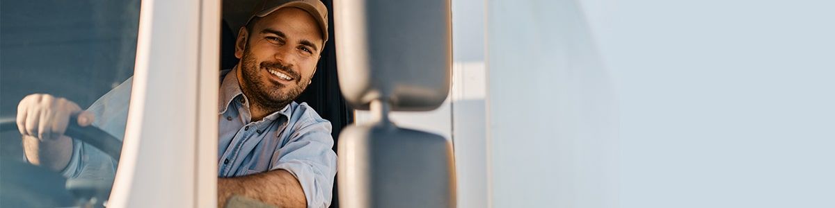 A trucker is smiling, wearing a tan cap and blue shirt. He's leaning out the window of his truck.