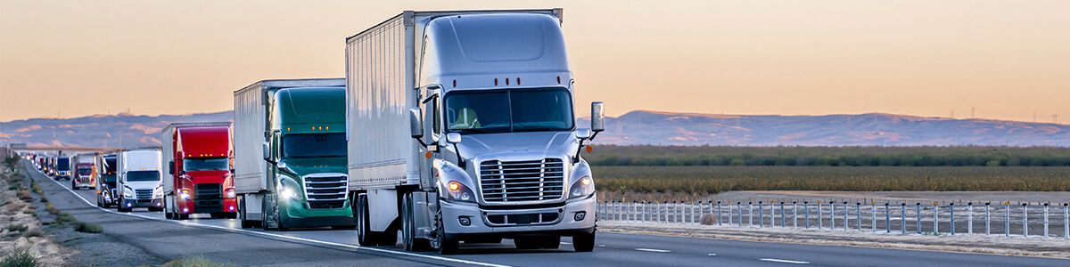 A line of multicolored semi trucks are driving down the highway. In the distance is a low mountain against the orange sky