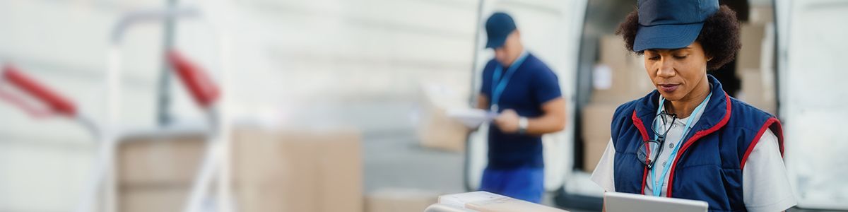 A woman wearing a blue, white, and red delivery uniform is holding a digital tablet and looking down at a brown box.