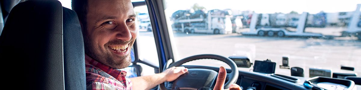 A trucker is sitting in the drivers seat, turning to smile at the person in the back of the cab taking the picture.